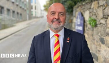 Man in his late 50s wearing a blue suit and a yellow and red striped tie. he is bald and has a white and grey beard