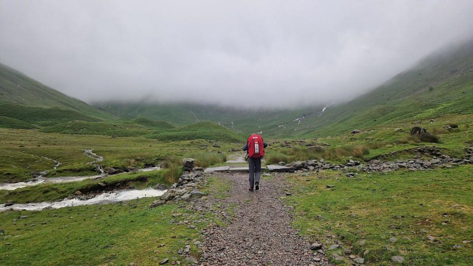 A mountain rescue volunteer, wearing a large red backpack, walks into the distance. There is a huge crag and fog in the distance, covering the Lake District fells.