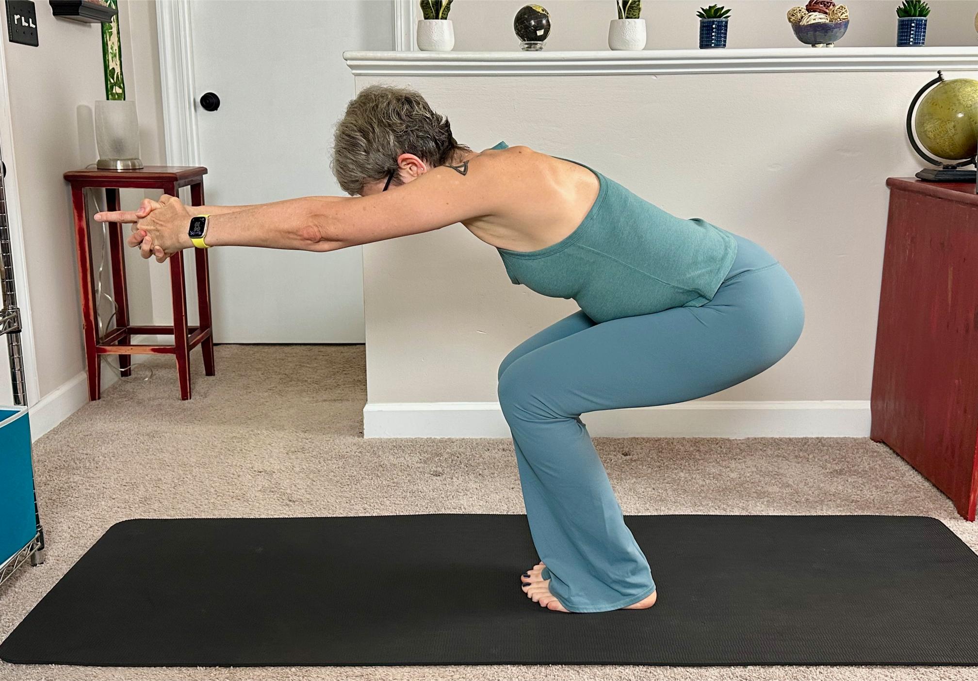 woman performing a chair pose in a living room setting on a black yoga mat wearing blue leggings and vest.