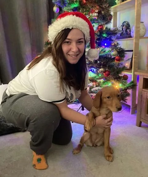 Woman in Santa hat kneeling with a puppy near a Christmas tree.