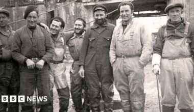 A group of seven whalers posing for the camera in a black and white picture. All are looking at the camera except the man in waders third from the left, who is staring at a man in overalls in a hat. The man on the near right is wearing waders and is leaning on a stick.