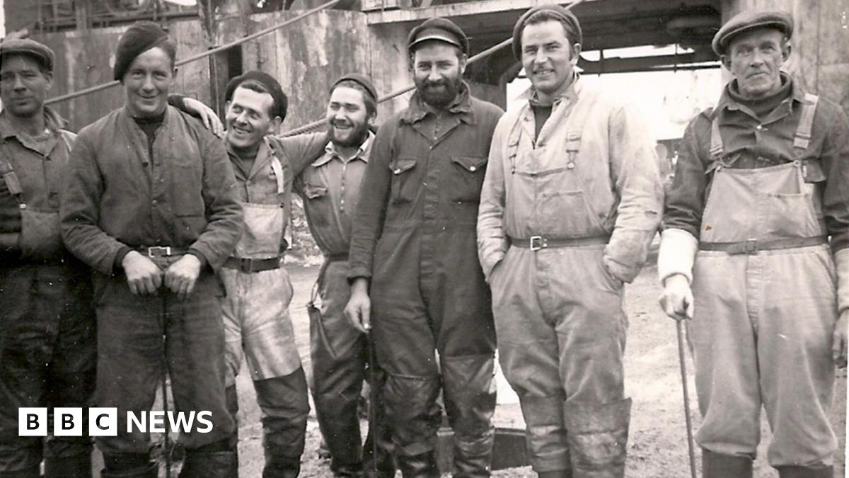 A group of seven whalers posing for the camera in a black and white picture. All are looking at the camera except the man in waders third from the left, who is staring at a man in overalls in a hat. The man on the near right is wearing waders and is leaning on a stick.