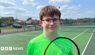 Oliver Appleby, 18, has brown hair with a fringe over his eyes. He has dark rimmed glasses and is wearing a lime coloured T-shirt. He is holding a tennis racquet. Behind him is the hard court.