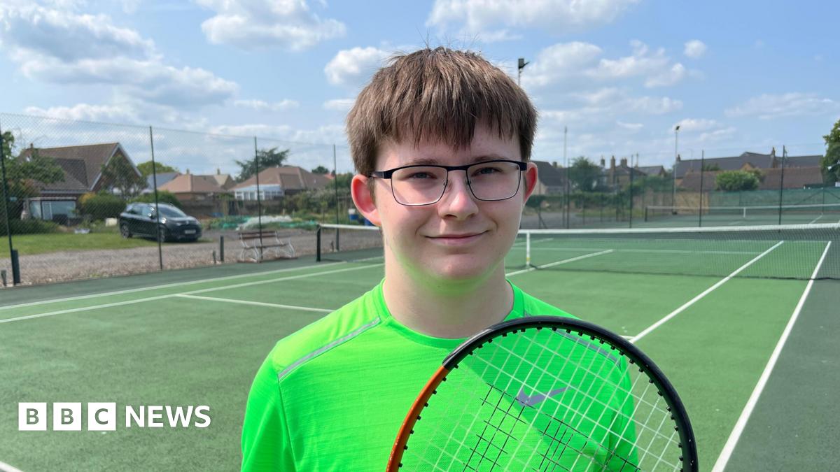 Oliver Appleby, 18, has brown hair with a fringe over his eyes. He has dark rimmed glasses and is wearing a lime coloured T-shirt. He is holding a tennis racquet. Behind him is the hard court.