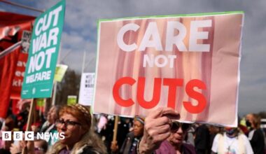 A crowd of people protesting, holding up signs including "care not cuts" and "cut war not welfare".