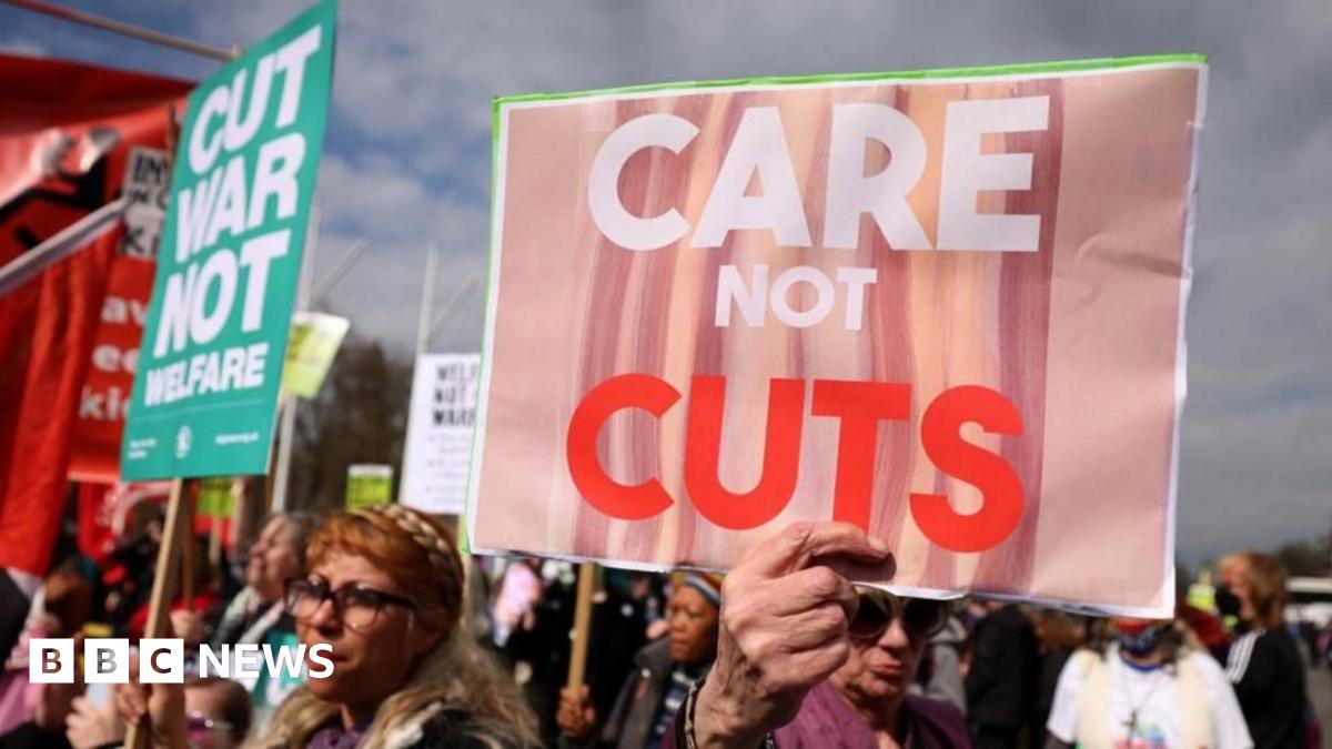 A crowd of people protesting, holding up signs including "care not cuts" and "cut war not welfare".