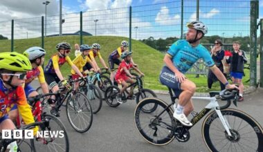 Mark Cavendish wears a blue jersey and a helmet and turns around on a bike and looks back at children who are following him.