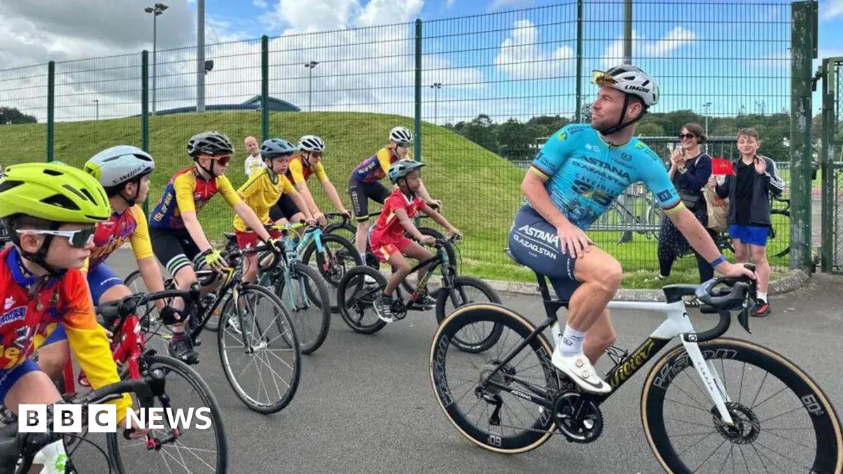 Mark Cavendish wears a blue jersey and a helmet and turns around on a bike and looks back at children who are following him.