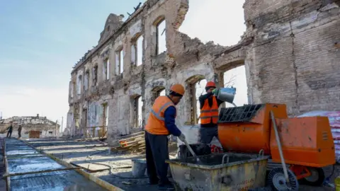 Getty Images Russian workers in Mariupol mix building materials. They are dressed in hard hats and orange high-vis jackets. Behind them is a war-damaged building with a chunk of its wall missing.