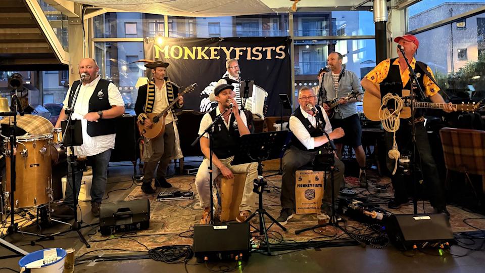 A band called Monkeyfists perform at the Portishead Sea Shanty Festival. They are inside an atrium-style space and are all in pirate fancy dress