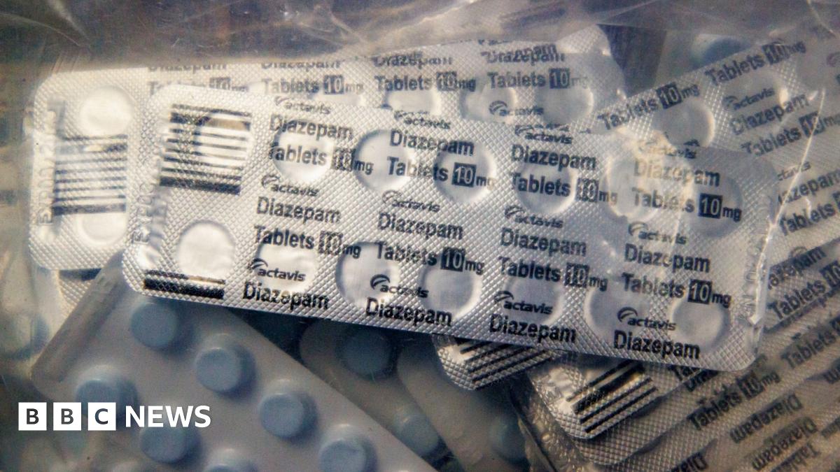 Large clear bags showing boxes of seized medicines. The bags are placed on the floor, with some placed on wooden desks around the room. There are also a few cardboard boxes which are also filled with white medicine boxes. There is a large poster in the background with 'Confronting Organised Crime through Agency Partnership' written on it.