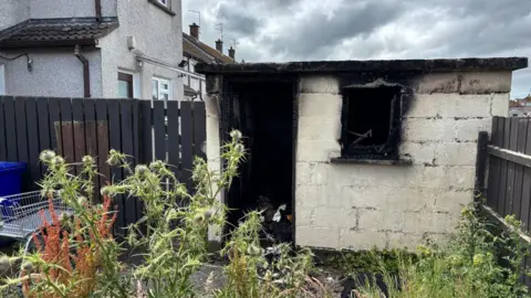 A concrete cream-coloured shed, with a burnt out window and door. There is a grey fence to the left of the shed and part of a grey house visible behind the fence. There are tall weeds growing in front of the shed. There is a cloudy sky above. 
