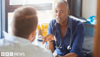 A GP and patient chat inside a treatment room. The GP is facing the patient who cannot be fully seen. The female GP has cropped grey hair and a blue shirt.