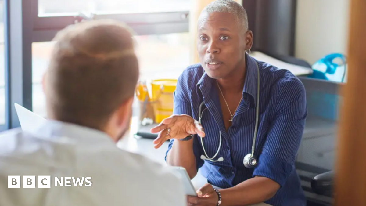 A GP and patient chat inside a treatment room. The GP is facing the patient who cannot be fully seen. The female GP has cropped grey hair and a blue shirt.