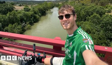 Mr White takes a selfie from his bike, which is positioned by some red railings on a bridge over a large river. He is wearing a green sports top and dark sunglasses.