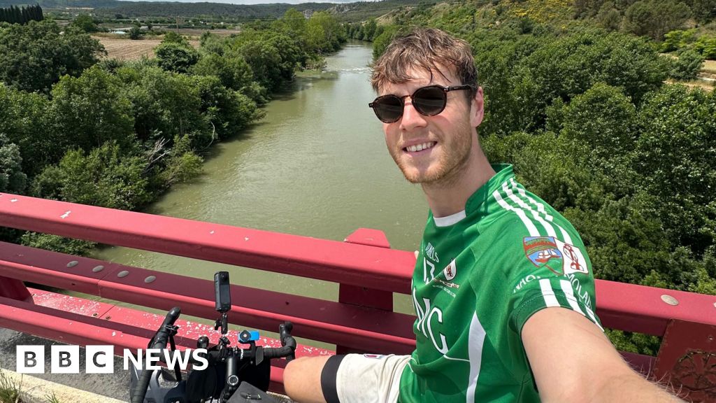 Mr White takes a selfie from his bike, which is positioned by some red railings on a bridge over a large river. He is wearing a green sports top and dark sunglasses.
