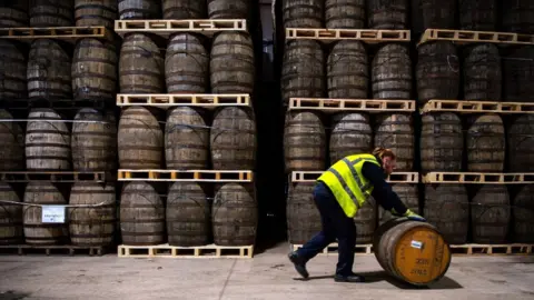 Getty Images A man wearing a high-visibility vest over dark clothes rolls a whisky cask along the ground. He is walking in front of a stack of whisky casks. 