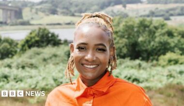 Vivian Tanaka smiles at the camera. She is on a hill that overlooks the River Orwell and Orwell Bridge near Ipswich. She wears a bright orange shirt and has blonde hair that has been braided and tied back.