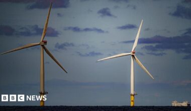 Two wind turbines next to each other in the sea. One is white and the other is slightly in shade. A ship can be seen to the right hand side.