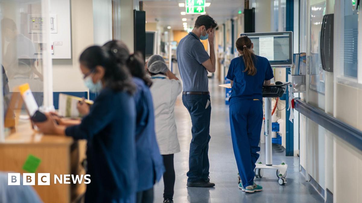 A hospital ward with several NHS workers dressed in navy blue or white uniforms at their posts.
