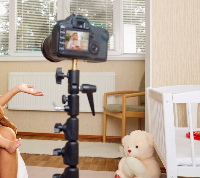 Adult sitting with a baby on a bed, looking at a camera on a tripod; they are surrounded by toys, including a teddy bear