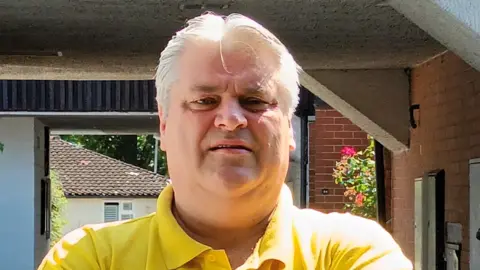 Stan Burridge A man with very blond hair and wearing a yellow polo top looking slightly down into the camera. There is a porchway in the background and a house beyond that.