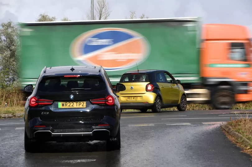 The image shows one car waiting at the junction, another car waiting in the central reservation, while a huge lorry drives at speed down the carriageway