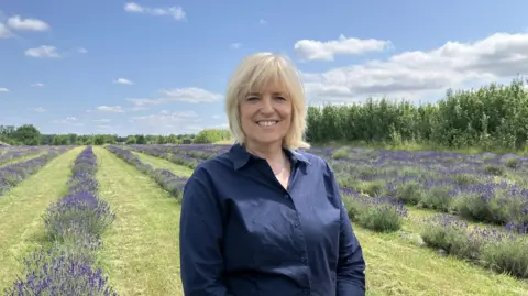 Emily Coady-Stemp/BBC A woman in a dark blue shirt stood in front of rows of planted lavender, separated by grass. The sky is bright and blue with a few clouds.