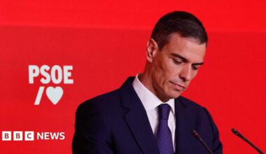 A man in a dark suit and tie looks down with a red backdrop