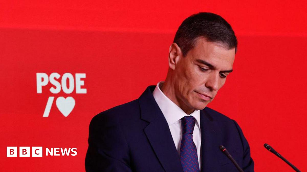 A man in a dark suit and tie looks down with a red backdrop