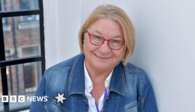 Rosemary Shrager stood in front of a white wall and black panelled window frame. She is wearing a blue denim jacket, white shirt and red-framed glasses.