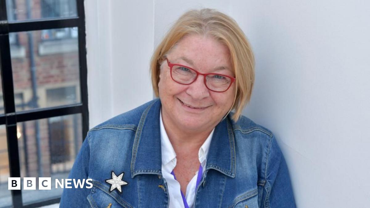 Rosemary Shrager stood in front of a white wall and black panelled window frame. She is wearing a blue denim jacket, white shirt and red-framed glasses.