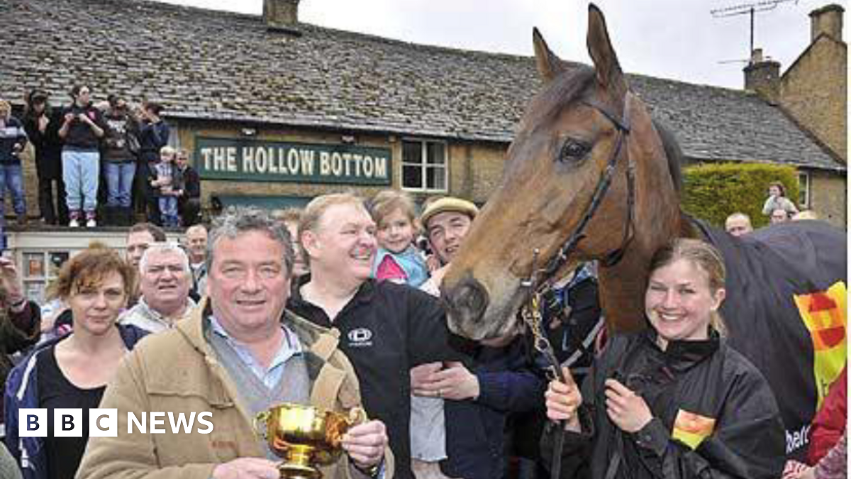 A large crowd stands outside of The Hollow Bottom - a traditional Cotswold pub - celebrating a Gold Cup win. People can be seen standing on a flat top roof above the door. One man at the front of the crowd holds a gold cup while a girl holds the winning horse's reins.