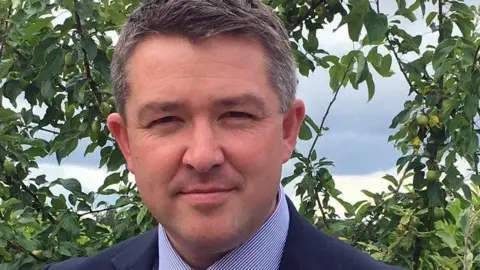 Herefordshire and Worcestershire ICB A man with short dark grey hair, wearing a dark blue suit and a blue pinstripe shirt, looks at the camera. He is standing in front of a fruit tree of some kind.
