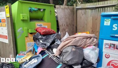 A pile of household waste, including black bags, full carrier bags and other items. The pile is between a large green recycling bin and a similar blue bin, and surrounded by wooden fencing.