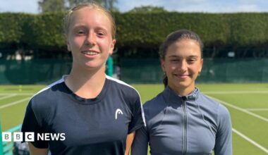 Daniella Britton (left) and Grace Watson stand between two tennis courts, smiling at the camera. Daniella has ginger hair and dental braces and wears a black T-shirt. Grace, who is standing slightly further back and appears to be the shorter of the two, has dark brown hair and wears a grey, zipped sports top.