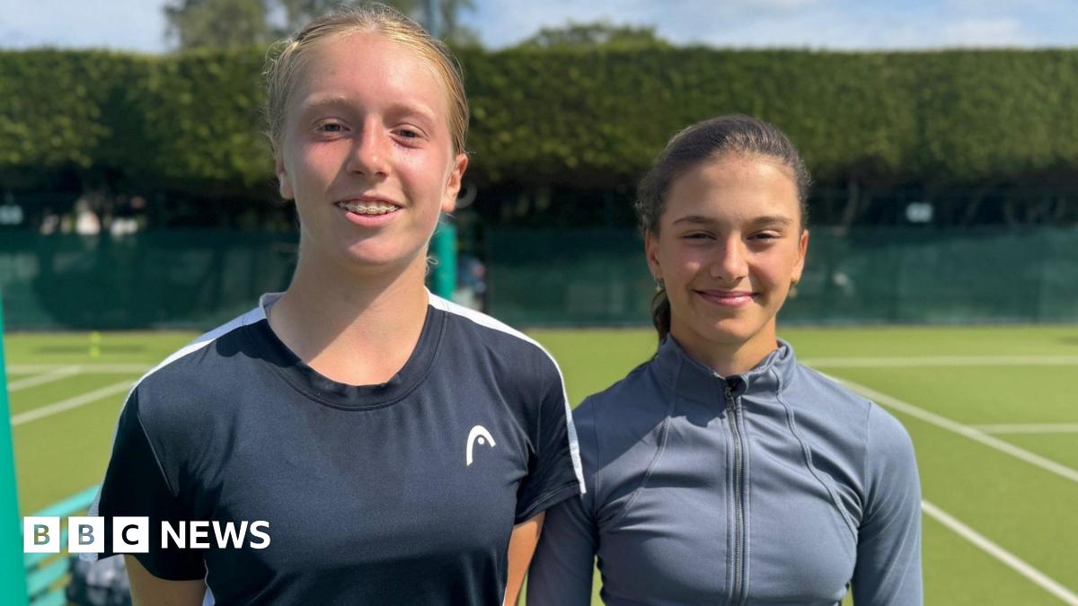 Daniella Britton (left) and Grace Watson stand between two tennis courts, smiling at the camera. Daniella has ginger hair and dental braces and wears a black T-shirt. Grace, who is standing slightly further back and appears to be the shorter of the two, has dark brown hair and wears a grey, zipped sports top.