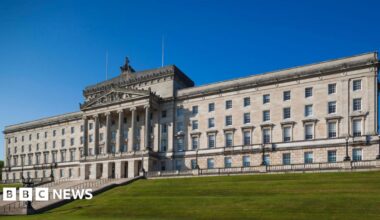A white washed grand Stormont with 365 windows and six pillars at the grand entrance. Behind the building is a blue sky and in front is steps and green grass.