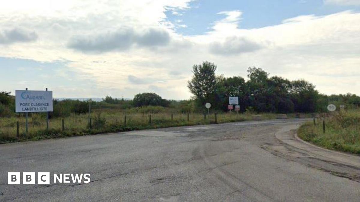 Entrance road leading to Augean at Port Clarence. There is a small white sign with the firm's name on it on the left-hand side of the road. The road is Tarmac and has a 30mph speed limit. It is surrounded by grassed areas and trees.