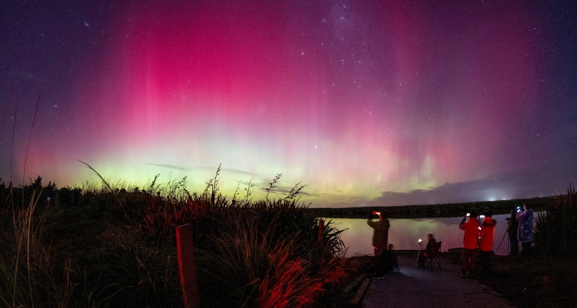 a group of people stand at the edge of a body of water to view bring yellow white and magenta waves of ionized gas in Earth