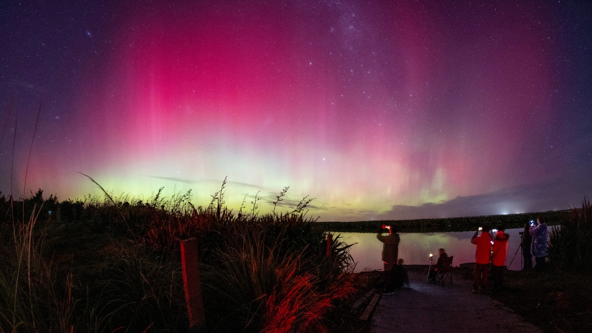 a group of people stand at the edge of a body of water to view bring yellow white and magenta waves of ionized gas in Earth's atmosphere, known as aurora australis or the southern lights