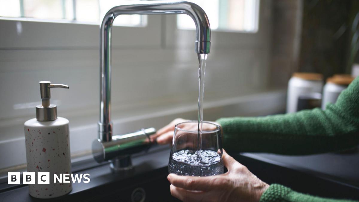 A woman is filling a glass with water from a silver tap. You can only see her hands. She is wearing a green cardigan.