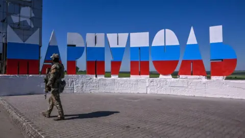 Getty Images A uniformed Russian soldier walks in front of a sign that spells "Mariupol" in Cyrillic, with the letters painted the colours of the Russian flag.