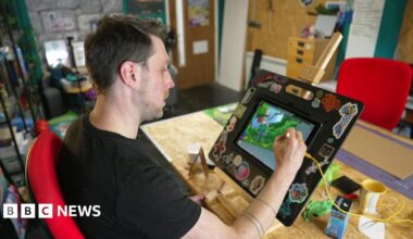 Nich Angell wearing a black t-shirt and sitting at a wooden desk in his art workshop. He is drawing a female comic book character on an iPad.