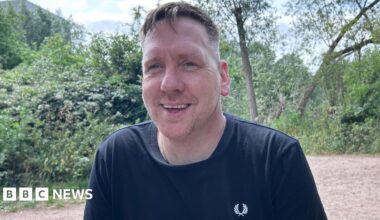 A man in a black Fred Perry T-shirt smiles at the camera against the backdrop of a green open space.