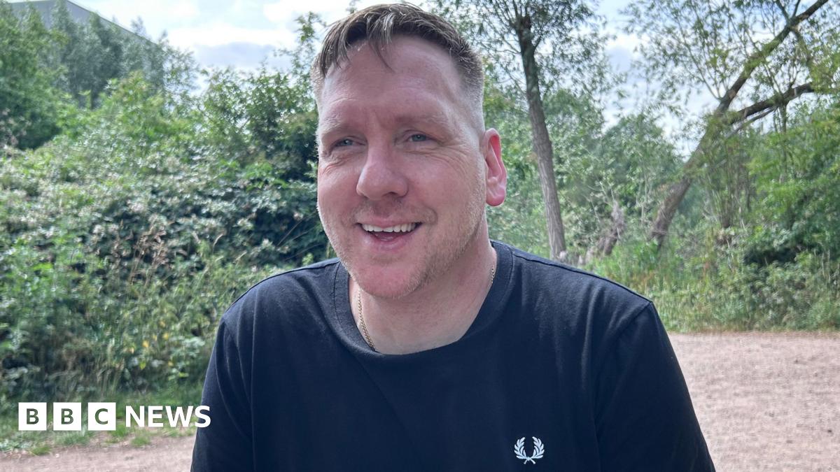 A man in a black Fred Perry T-shirt smiles at the camera against the backdrop of a green open space.