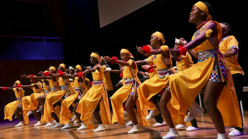 A group of dozens of young woman who are part of the touring Project Zulu Choir dance on stage at Bristol Beacon. They are wearing yellow headbands and dresses with multicoloured shorts underneath