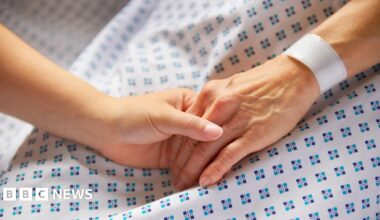 Woman in medical gown with wristband on. Her hand is held by another woman.
