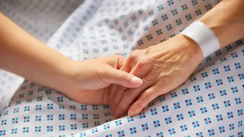 Getty Images Woman in medical gown with wristband on. Her hand is held by another woman. 