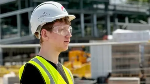 BBC Blake Smith-Helps wearing hi-vis, a helmet and protective glasses on a construction site. 